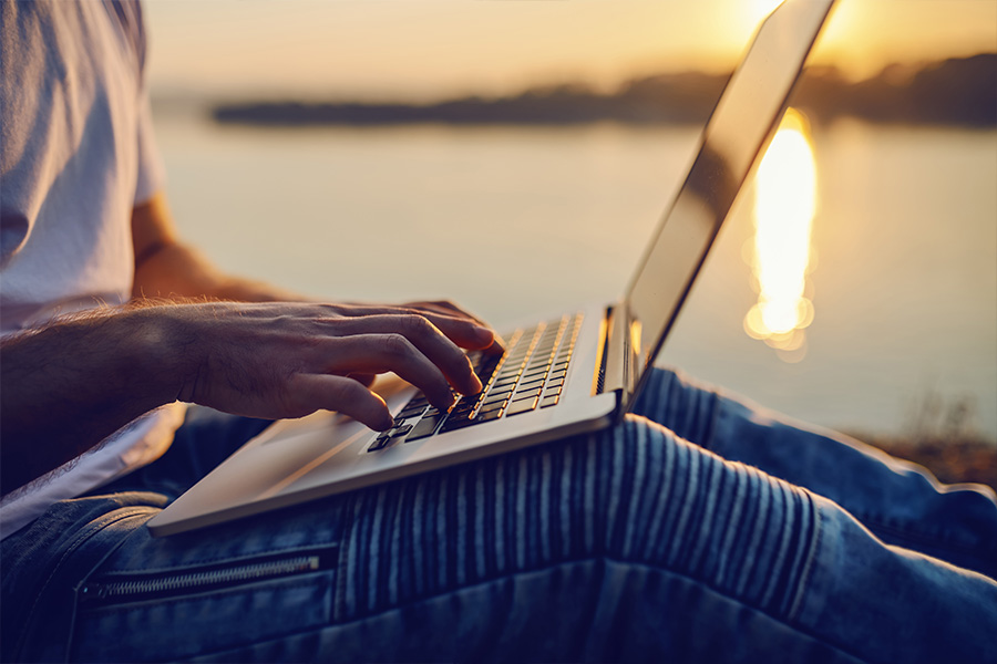 Blog - Close Up of Person Sitting on the Ground Next to a Bay While Typing on a Laptop During Sunset